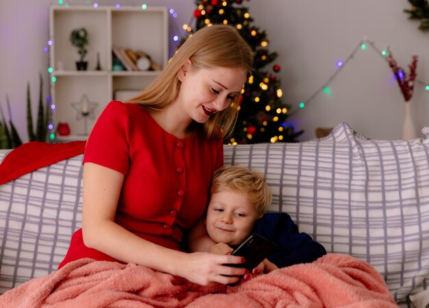 Счастливая мать в красном платье сидит на диване со своим маленьким ребенком под одеялом со смартфоном в украшенной комнате с елкой в стене
