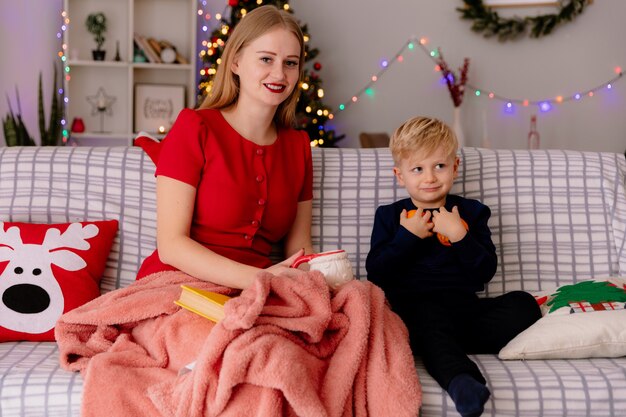 Happy mother in red dress with cup of tea and her little child  holding oranges under blanket in a decorated room with christmas tree in the background