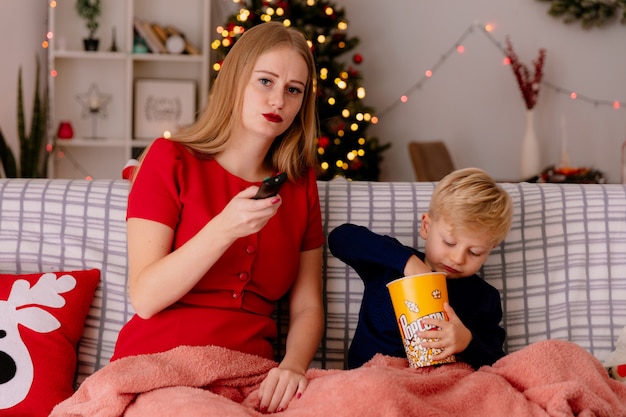 Счастливая мать в красном платье со своим маленьким ребенком, сидящим на диване под одеялом с ведром попкорна, вместе смотрит телевизор в украшенной комнате с рождественской елкой в стене