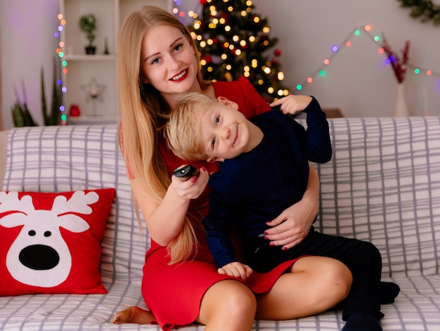 Felice madre in abito rosso con il suo bambino seduto su un divano che si diverte a guardare la tv insieme in una stanza decorata con albero di natale nel muro