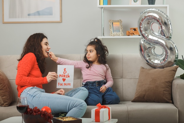 Happy mother with her little child daughter sitting on a couch holding greeting card smiling cheerfully in light living room celebrating international women's day march 8