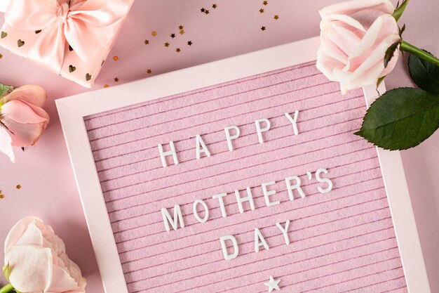 Happy mothers day words on pink felt letter board. Festive composition with roses and a box with a gift on a pink surface. Top view, flat lay. Copy space.