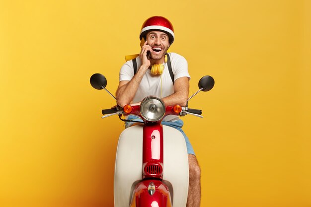 Happy motorcyclist poses on fast own transport, calls client via smartphone, travels on long distance, wears helmet, stereo headphones around neck, smiles at camera. Male driver drives scooter