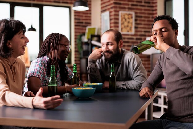 Happy multiracial friends sitting at home in living room while talking and enjoying snacks and beverages. Cheerful young adults having a conversation while relaxing with positive lifestyle.