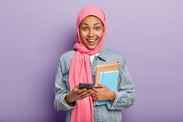 技術の幸せなイスラム教徒の女性の高度なユーザーは、メモ帳と携帯電話を保持し、頭にピンクのベールを身に着けています