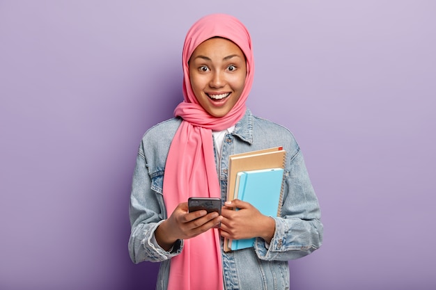 Happy Muslim woman advanced user of technology holds notepads and cellular, wears pink veil on head