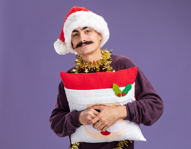 보라색 벽 위에 서있는 얼굴에 미소로 크리스마스 베개를 들고 그의 목 주위에 반짝이와 크리스마스 산타 모자를 쓰고 행복 콧수염 남자