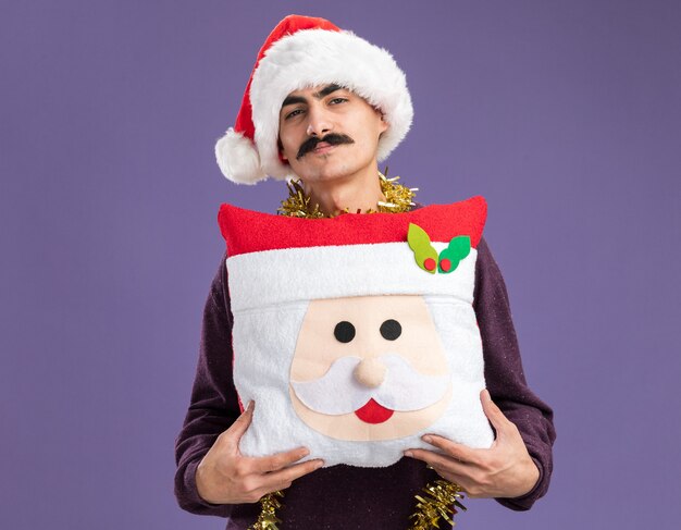 Free photo happy mustachioed man wearing christmas santa hat  with tinsel around his neck holding christmas pillow  with smile on face standing over purple wall