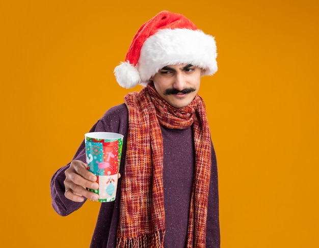 Happy mustachioed man wearing christmas santa hat with warm scarf around his neck holding colorful paper cup looking at it with smile on face standing over orange background