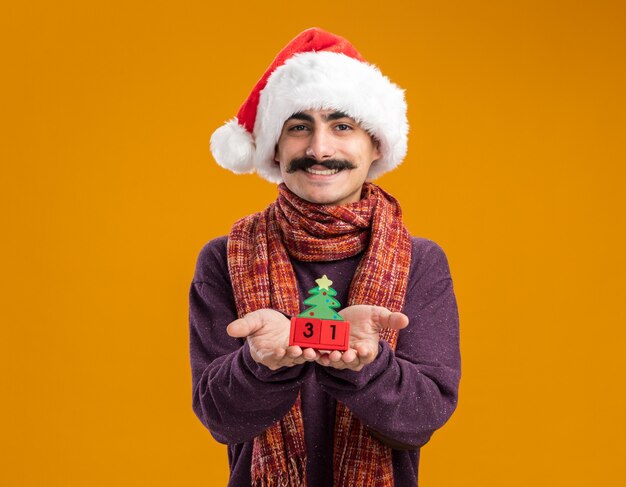 Happy  mustachioed man wearing christmas santa hat  with warm scarf around his neck showing toy cubes with new year date smiling cheerfully  standing over orange wall