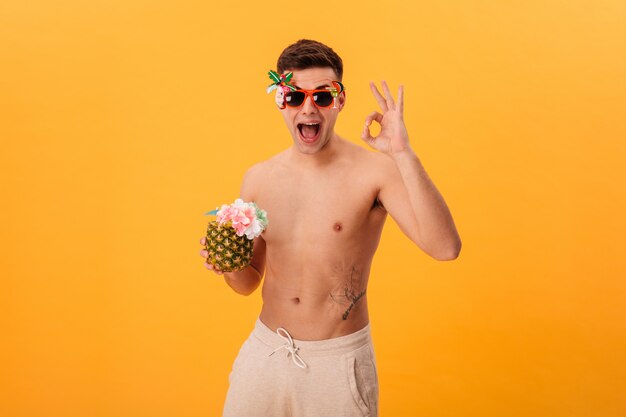 Happy naked man in shorts and unusual sunglasses holding cocktail while showing ok sign 
