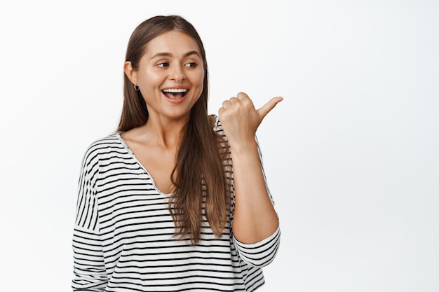 Happy natural girl laughing, smiling and pointing finger right, looking at logo, sale advertisement aside, white background. Copy space