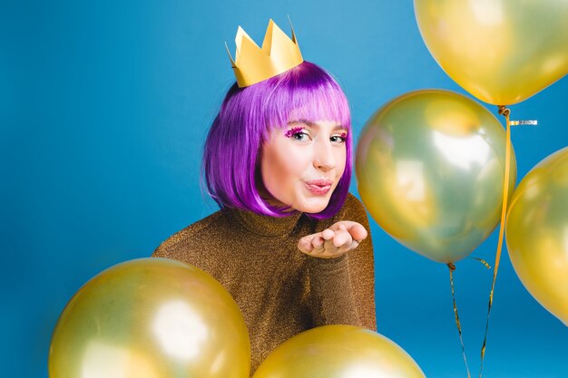 Happy new year party time of attractive young woman sending a kiss, surround golden balloons . Cut purple hair, luxury dress, having fun, birthday celebration.