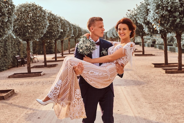 Happy newlyweds. Handsome groom holds his charming bride in her arms in the amazing garden.