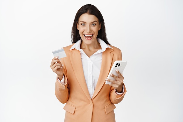 Happy office lady in suit holding mobile phone and credit card shopping online purchase in application on smartphone standing over white background