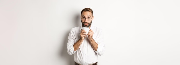 ホット コーヒーを飲み、白い背景の上に立ってうわさ話を興奮して見て幸せなオフィス ワーカー