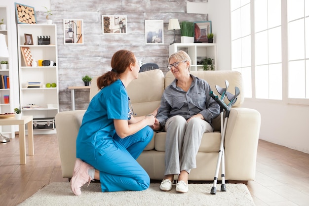 Happy old woman in a nursing home sitting on couch talking with her caretaker. Retired woman with crutches.