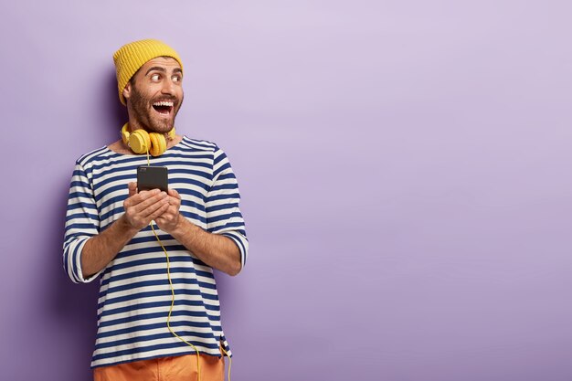 Счастливый оптимистичный молодой человек смотрит в сторону, держит современный мобильный телефон, просматривает музыкальную платформу в Интернете, загружает песню в плейлист, носит желтые наушники на шее