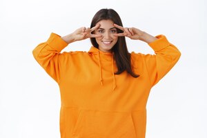 Happy optimistic caucasian female in orange hipster hoodie show goodwill peace gesture over eyes disco kawaii signs and smiling joyfully enjoy party having fun with friends take photo