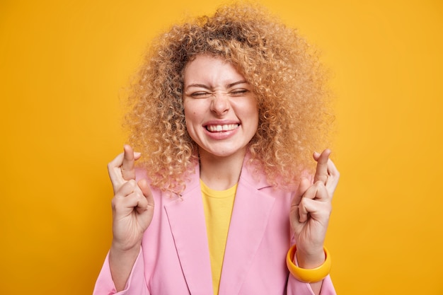 Happy optimistic woman with natural curly hair waits for fortune crosses fingers grins at camera