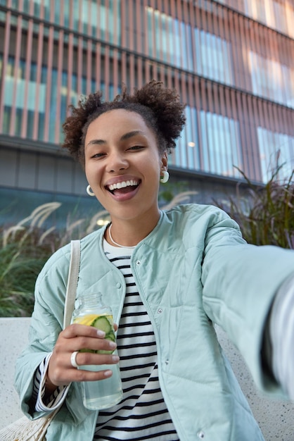 Free photo happy optimistic woman with two hair buns dressed in jacket enjoys free time and walking in city holds bottle of detox drink