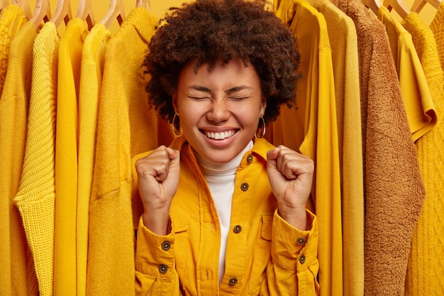 Happy overjoyed dark skinned woman stands near yellow stylish clothes on hangers, clenches fists, rejoices successful purchase