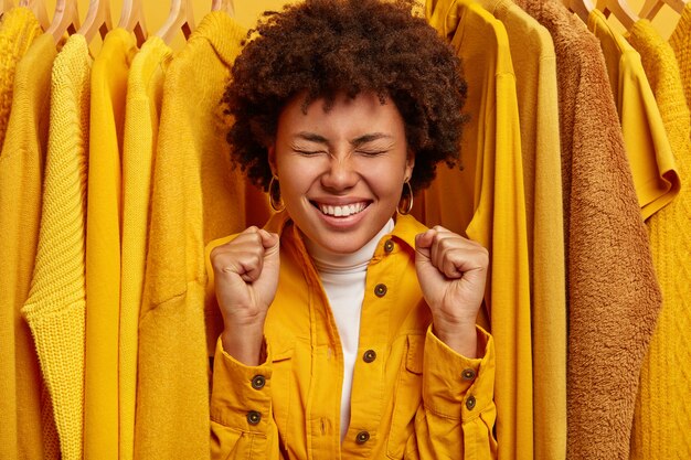 Happy overjoyed dark skinned woman stands near yellow stylish clothes on hangers, clenches fists, rejoices successful purchase