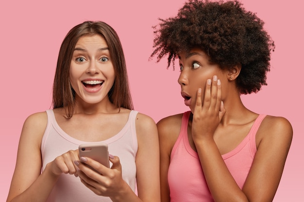 Happy overjoyed female model shows text message on cellular to her dark skinned friend who has surprised expression