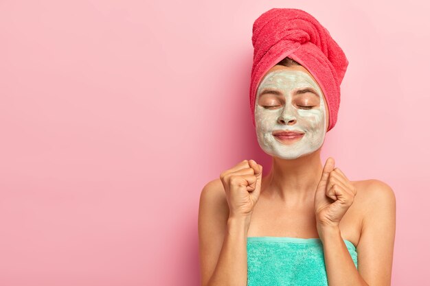 Happy overjoyed woman applies facial clay mask, has rejuvenation treatment, clenches both fists, wears towel, keeps eyes closed, isolated on pink studio wall