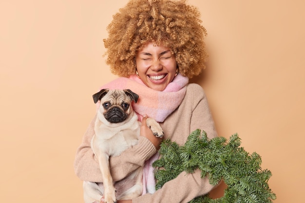 Free photo happy overjoyed woman bought green spurce wreath holds pedigree dog has festive mood on christmas eve wears sweater and scarf around neck isolated over beige background. people pets holidays