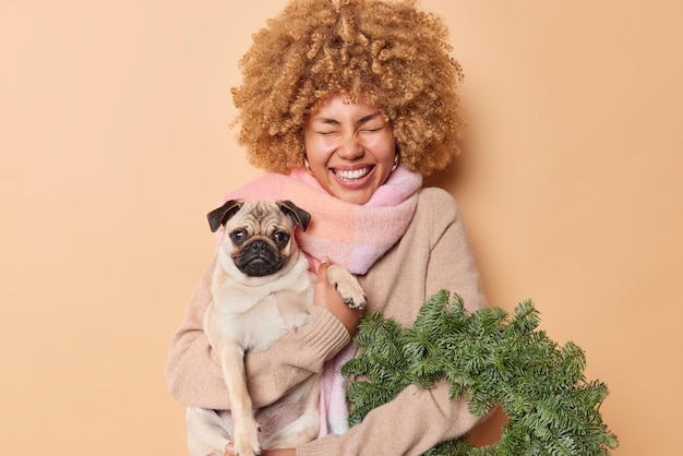 Felice donna felicissima ha acquistato la corona di spurce verde tiene il cane di razza ha un'atmosfera festosa alla vigilia di natale indossa maglione e sciarpa intorno al collo isolato su sfondo beige. persone animali domestici vacanze