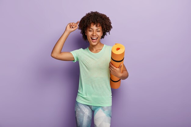 Happy overjoyed woman holds yoga mat, raises arm, dressed in casual t shirt and leggings, being pleased by active workout