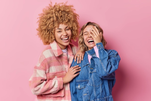 Happy overjoyed young women laugh gladfully hear something very funny dressed in fashionable clothes stand closely to each other isolated over pink background Friendship and emotions concept