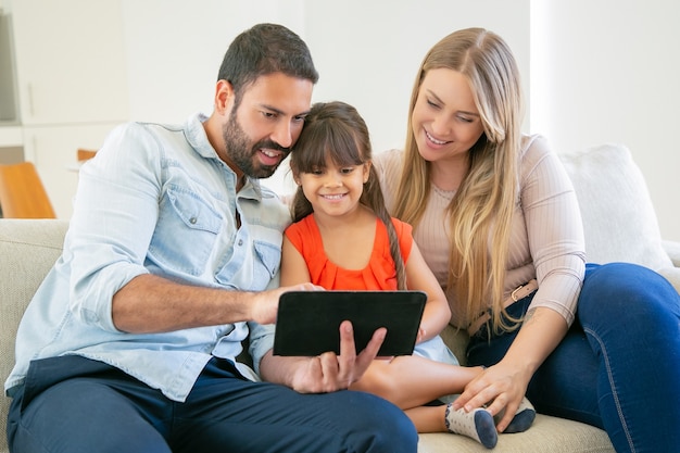 Happy parents and cute daughter sitting on couch, using tablet for video call or movie watching.