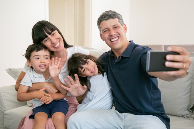 Happy parents hugging adorable kids, sitting on couch at home together, taking selfie on phone