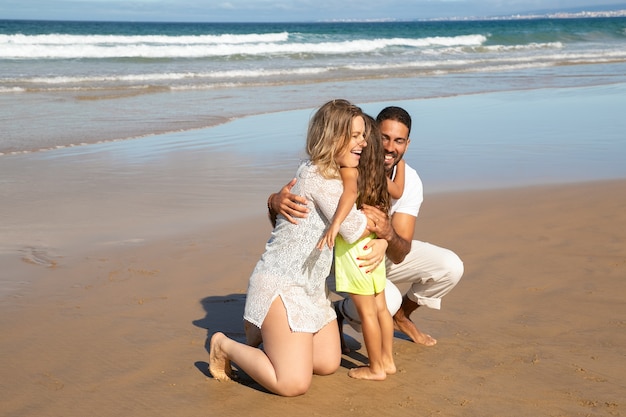 Happy parents hugging little daughter on wet sand at sea