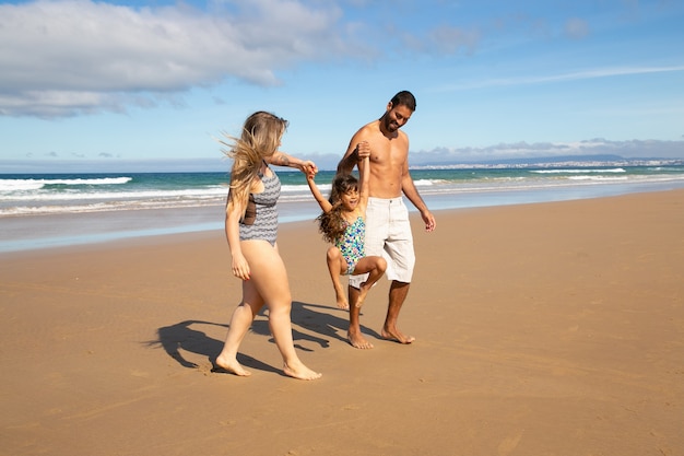 Happy parents and little girl wearing swimsuits, walking on golden sand from sea