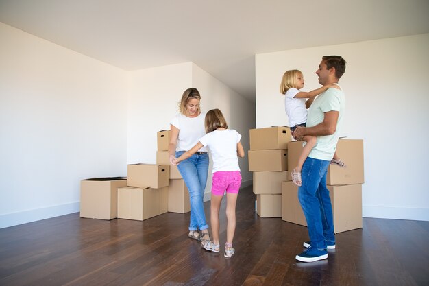 Happy parents and two girls dancing and having fun near heaps of boxes while moving into new flat