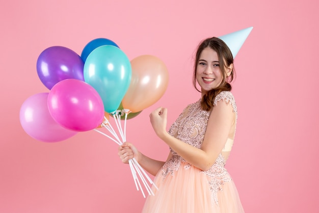 Ragazza felice festa con tappo di partito che tiene palloncini sul rosa