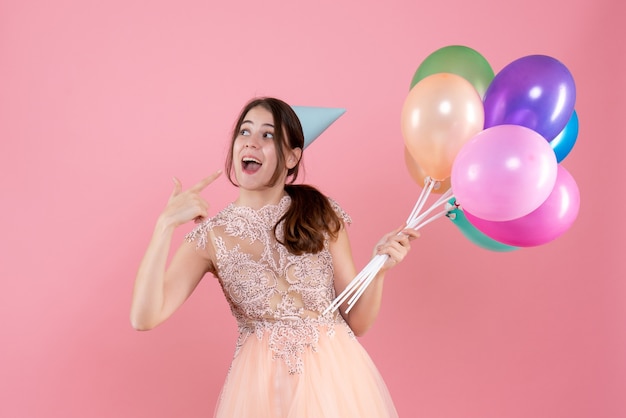 happy party girl with party cap holding balloons pointing with finger her smile on pink