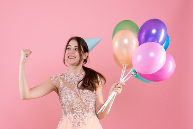 happy party girl with party cap holding balloons showing her muscle on pink