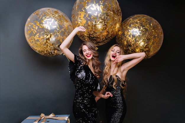 Happy party moments of two fashionable funny young women. Luxury black dress, red lips, long curly hair, brightful mood, having fun, big balloons with golden tinsels.