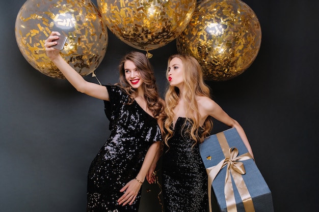 Momenti di festa felice di due giovani donne alla moda che fanno selfie. abito nero di lusso, lunghi capelli ricci, grandi palloncini con orpelli dorati, presente, divertirsi, sorridere.