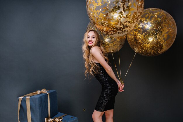 Happy party time amazing joyful young woman in black luxury dress, with long curly blonde hair holding big balloons full with golden tinsels. Presents, smiling, great mood.