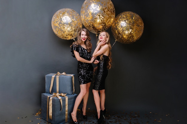 Счастливое времяпровождение двух очаровательных молодых женщин в роскошных черных платьях. Длинные вьющиеся волосы, привлекательный вид, подарки, большие шары с золотой мишурой, улыбка, веселье.