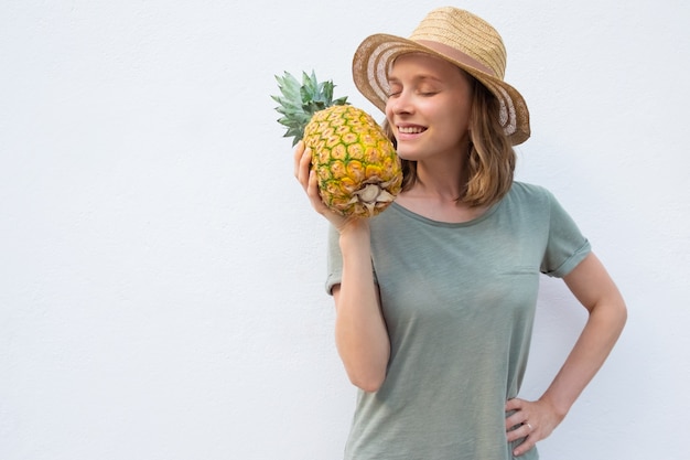 Happy peaceful woman in summer hat smelling whole pineapple