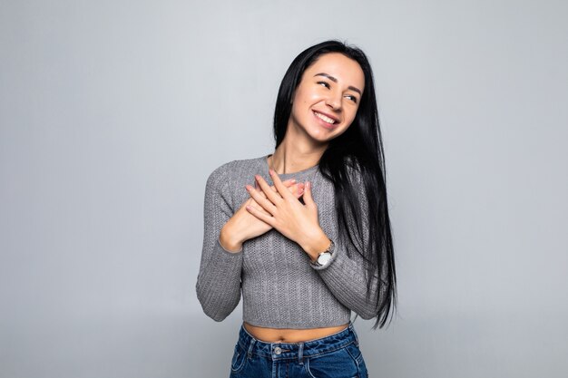 Happy peaceful womanisolated on grey studio wall hold hands at chest close to heart, smiling grateful female stand feel thankful, show appreciation and love