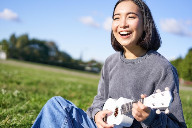 Happy people and hobbies smiling asian girl playing ukulele guitar and singing sitting in park outdo