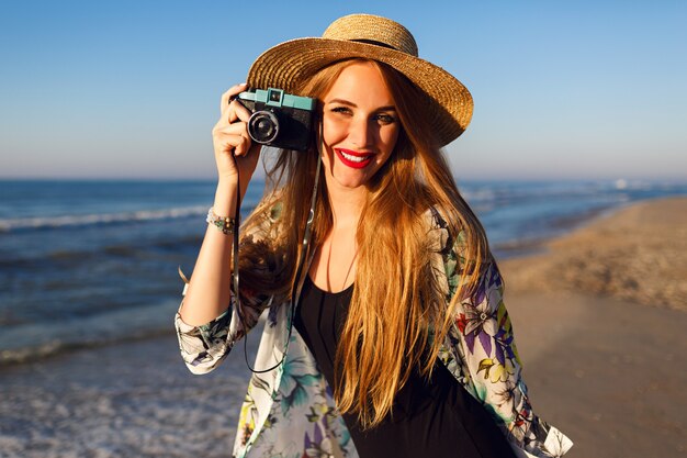 Happy petty woman with long blonde hairs having fun and making pictures on the beach near ocean on vintage camera, sunny colors