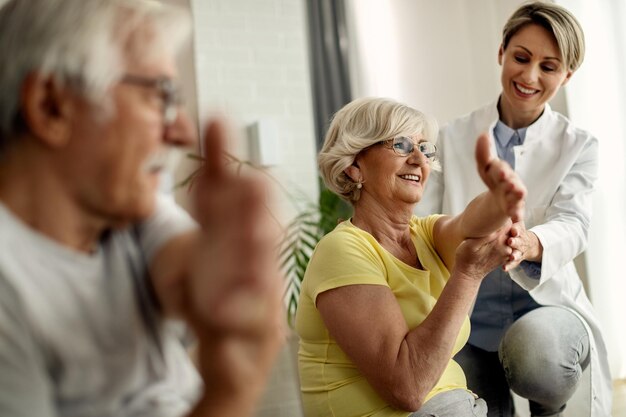 Happy physiotherapist helping to senior couple with stretching exercises at home
